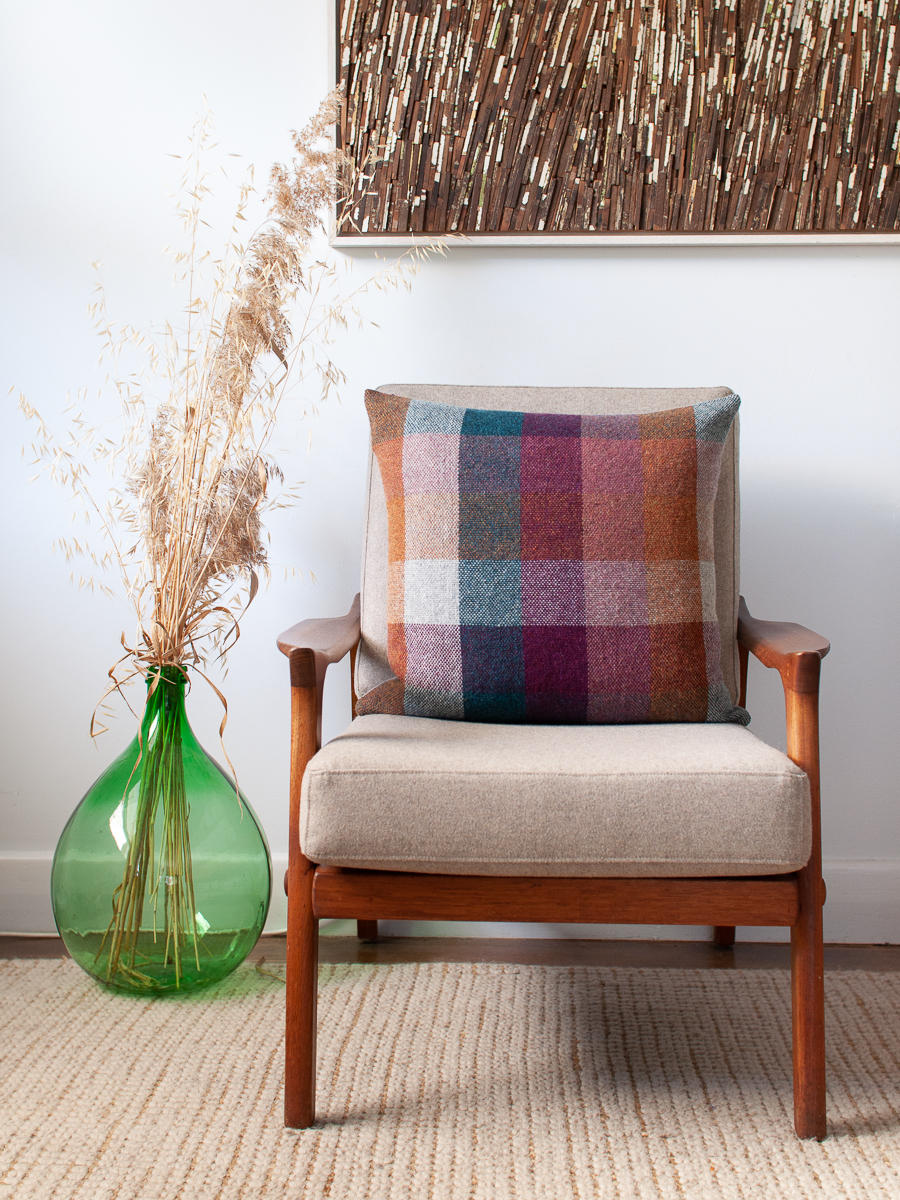 Handwoven cushion cover made with a 5 colour 3 inch check pattern on the front in shades of orange, dirty pink, bright purple, teal and silver grey. One Teal check is bordered with a playful short fringe on all 4 sides. The cushion is resting on a mid century danish deluxe chair next to an antique green demijohn filled with dry grasses.
