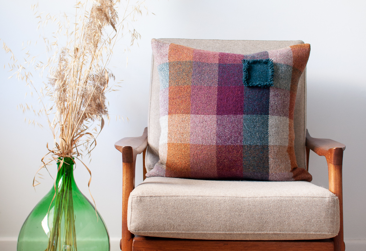 Handwoven cushion cover made with a 5 colour 3 inch check pattern on the front in shades of orange, dirty pink, bright purple, teal and silver grey. One Teal check is bordered with a playful short fringe on all 4 sides.