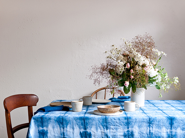A shibori dyed indigo tablecloth adorns a table with handmade pottery plates and vessels. A large vase of flowers is set to the right of the centre frame.